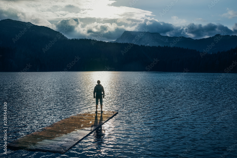 Silhouette man standing on the wooden raft on the blue lake. Cloudy mountain in the back with the evening light in nature landscape. Reflexion on the raft. Black Lake, Montenegro, Durmitor