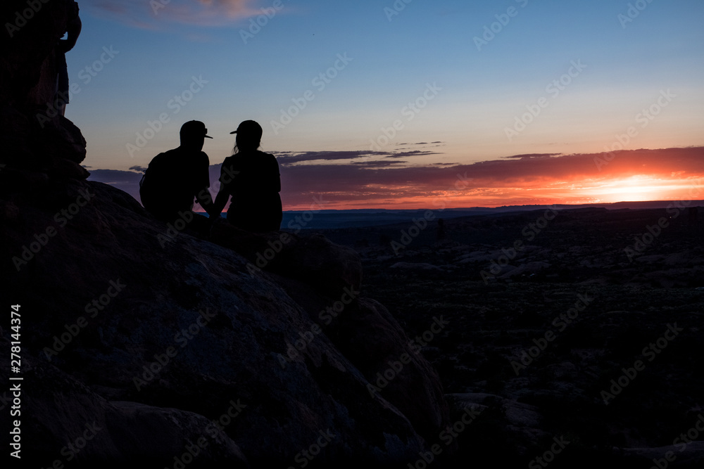 Couple of romantic tourists sitting on rocks watching sunset - Arches National Park, Utah
