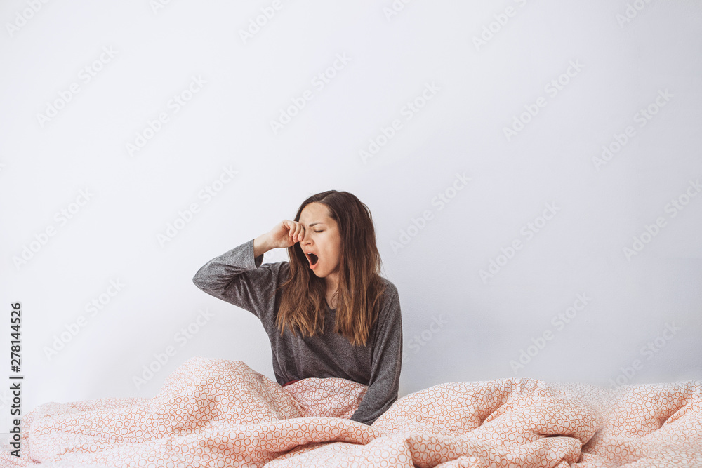 The girl is in bed and yawns. She covers her mouth with her hand. Early morning and she is sleepy.
