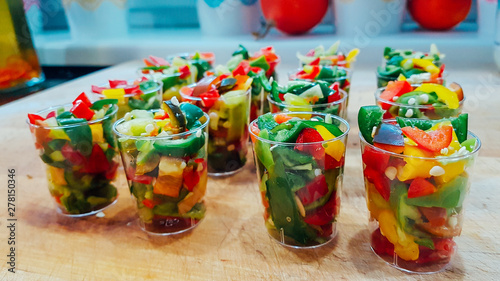 Chopped chili pepper pieces stored in plastic shot glasses. Pieces of paper are green, yellow and red. Some seeds in between them. Cups are placed on a wooden board. Spicy looking vegetable.