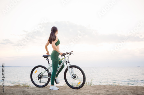 Beautiful young brunette woman resting after bicycle ride at sunrise. Model listening to music with wireless earbuds in the morning. © ianachyrva