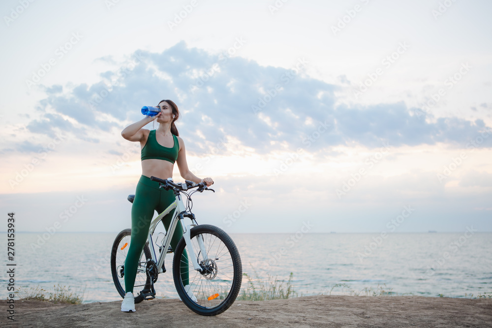 Young brunette woman with wireless earbuds and smart watches resting after bicycle ride at sunrise drinking water.