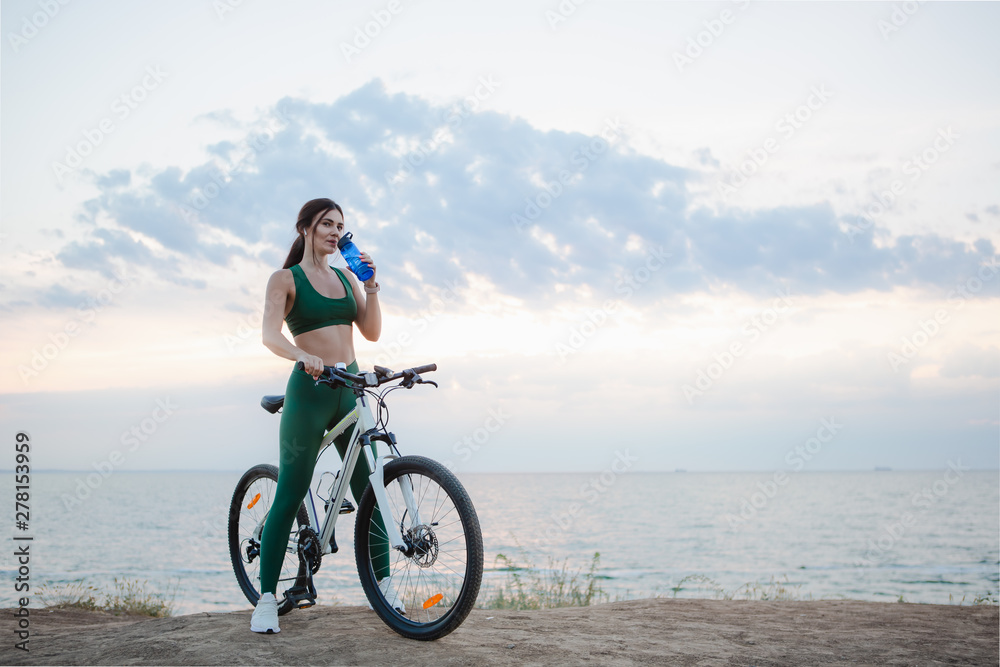 Young brunette woman with wireless earbuds and smart watches resting after bicycle ride at sunrise drinking water. Young brunette woman with wireless earbuds and smart watches resting after bicycle ride at sunrise drinking water.