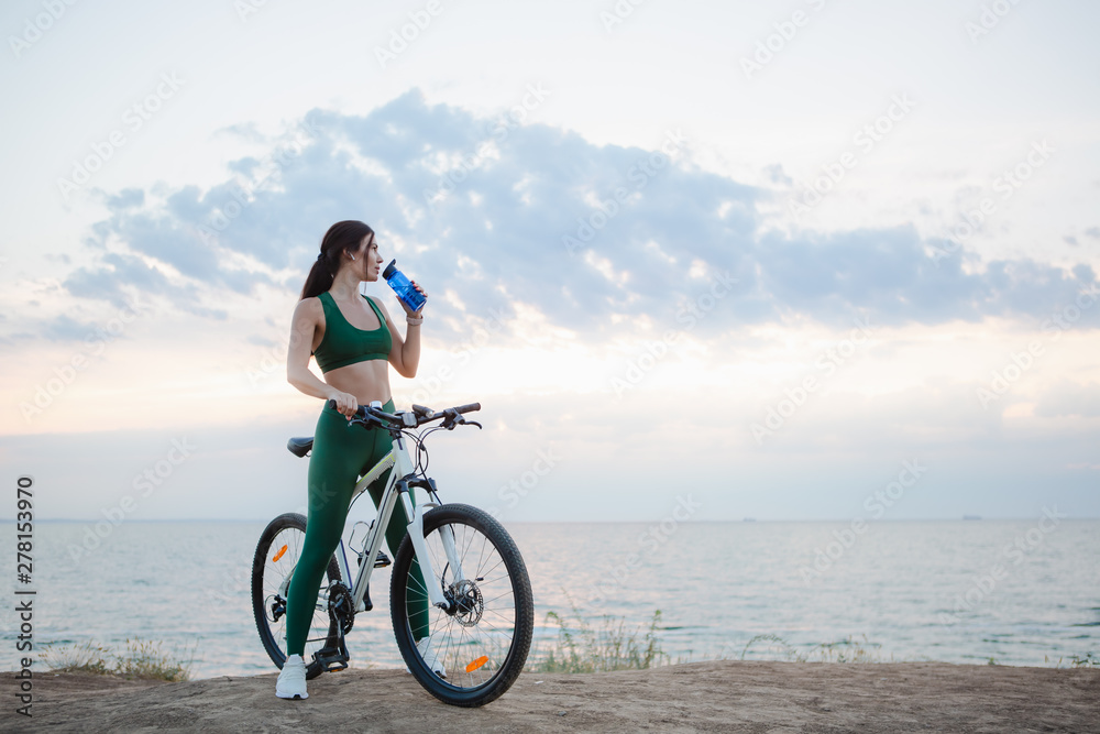 Young brunette woman with wireless earbuds and smart watches resting after bicycle ride at sunrise drinking water. Young brunette woman with wireless earbuds and smart watches resting after bicycle ride at sunrise drinking water.