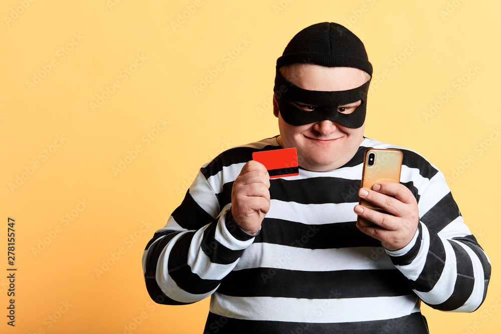 happy cheerful plump hacker with a bank card and mobile phone Isolated on the yellow Background. crime, illegal business copy space, isolated yellow background