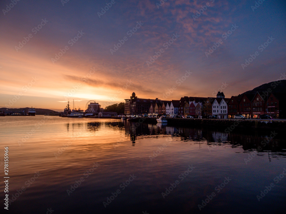 Beautiful front line of houses build in the port in Bergen, Norway. Soft reflections of houses and boats in the sea. Behind the buildings there is a small hill. Golden hour, sunset in the port.