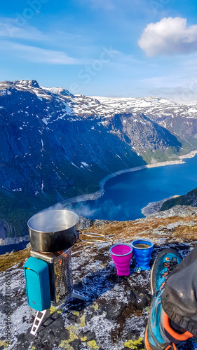 A camping stove with a pot with boiling water on it, with a view on Ringedalsvatnet lake, Norway. Next to it a hiking shoe and two cups. Slopes of the mountains are partially covered with snow. photo