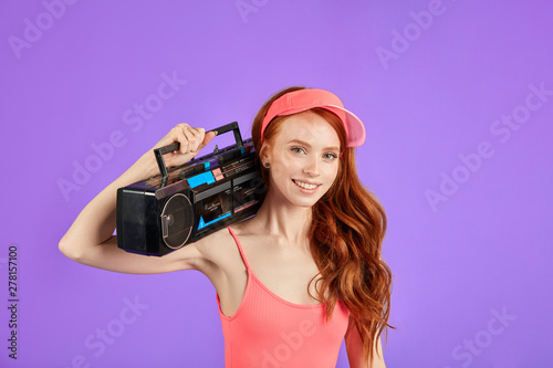 red-haired young woman with freckles looks in camera with smile, holds stereo and cassette tape player on her shoulder, wears pink sun cap and aerobics bodysuit photo