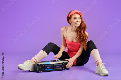 girl with nice freckles expresses positive sincere emotions, being amused friend, dressed in comfortable 80s style aerobics outwear, presses play button on portable retro audio player, sits on floor photo