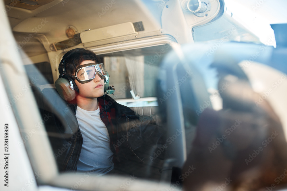 portrait of cute little boy made through the window of light single-engine airplane, dressed in large leather pilot jacket, pilot headset and aviator glasses, looking thoughtfully in sky.