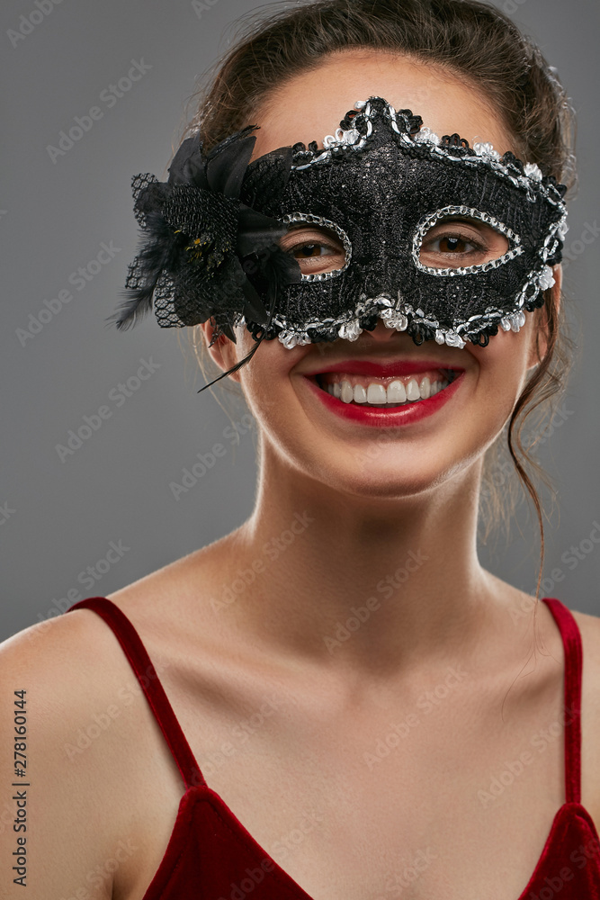Portrait of laughing lady with tied back brunette hair, wearing wine red crop top. The girl is looking at camera, wearing ink black carnival mask, adorned with massive flower, feathers and glitter. Portrait of laughing lady with tied back brunette hair, wearing wine red crop top. The girl is looking at camera, wearing ink black carnival mask, adorned with massive flower, feathers and glitter.