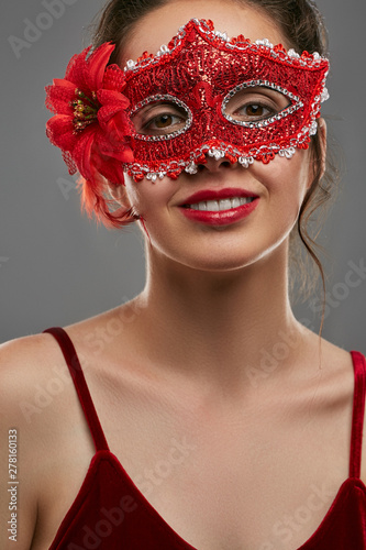 Fototapeta Naklejka Na Ścianę i Meble -  Portrait of smiling lady with tied back brunette hair, wearing wine red crop top. The girl is looking at camera, wearing rich red carnival mask, adorned with massive flower, feathers and glitter.