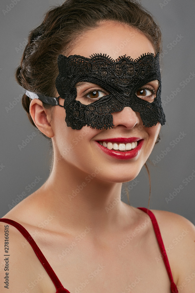 Half-turn shot of girl with dark hair, wearing wine red crop top. The smiling lady is looking at camera, wearing black carnival mask with perforation and rounded edges. Vintage carnival accessory.