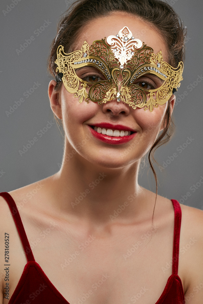 Half-turn shot of girl with dark hair, wearing wine red crop top. The  smiling lady is looking at the camera, wearing golden Venetian carnival  mask with fancy perforation. Vintage carnival accessory. Stock