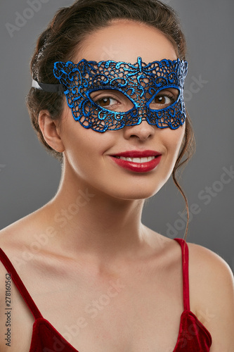 Portrait of girl with tied back brunette hair, wearing wine red crop top. The lady with slight smile is looking at camera, wearing blue carnival mask with perforation. Vintage carnival accessory.