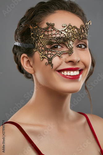 Half-turn shot of smiling lady, wearing wine red crop top. The woman is looking at the camera, wearing asymmetric golden carnival mask with perforation in view of spider web, tied with satin ribbon. photo