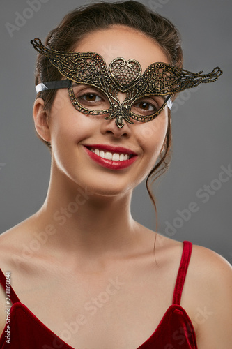 Half-turn portrait of smiling woman with dark hair, wearing wine red crop top. The young girl is tilting her head, wearing golden carnival mask with fancy perforation in view of heart in forehead. photo