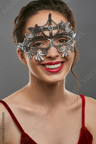 Half-turn shot of smiling lady, wearing wine red crop top. The young woman is tilting head, wearing asymmetric silver carnival mask with perforation and cross on forehead, tied with satin ribbon. photo