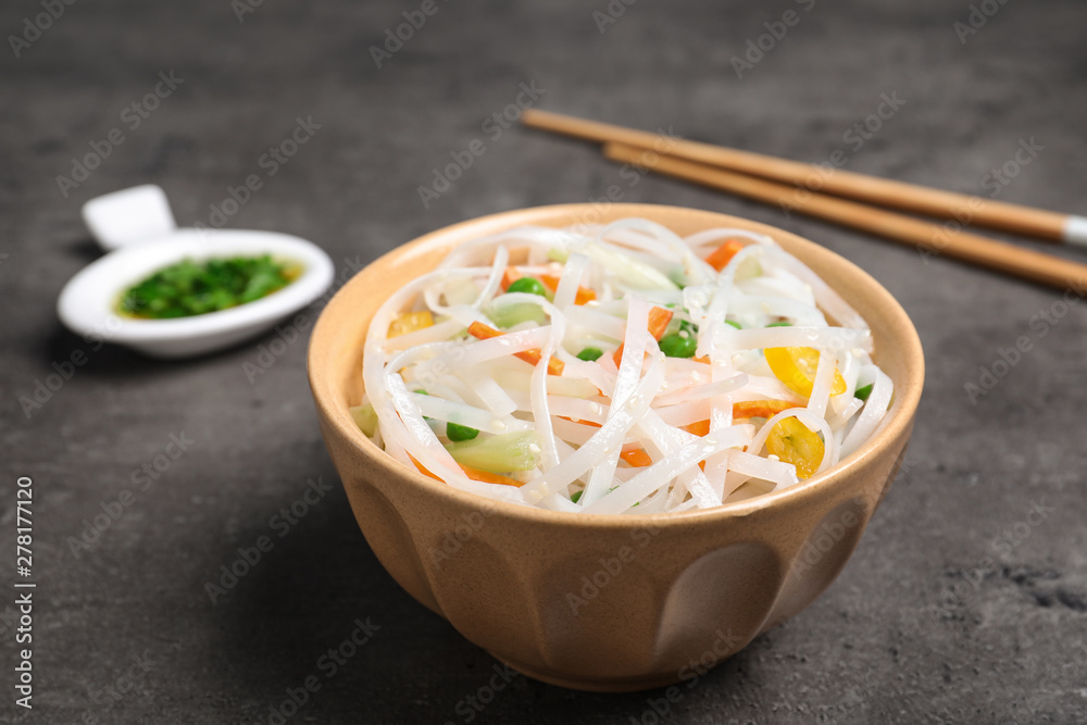 Bowl of rice noodles with vegetables and chopsticks on grey background