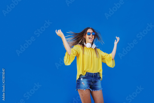 Young cheerful Caucasian teenage girl in yellow blouse and denim shorts jumping in front of blue background.