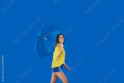 Beautiful smiling Caucasian teenage girl in yellow blouse walking with blue umbrella.