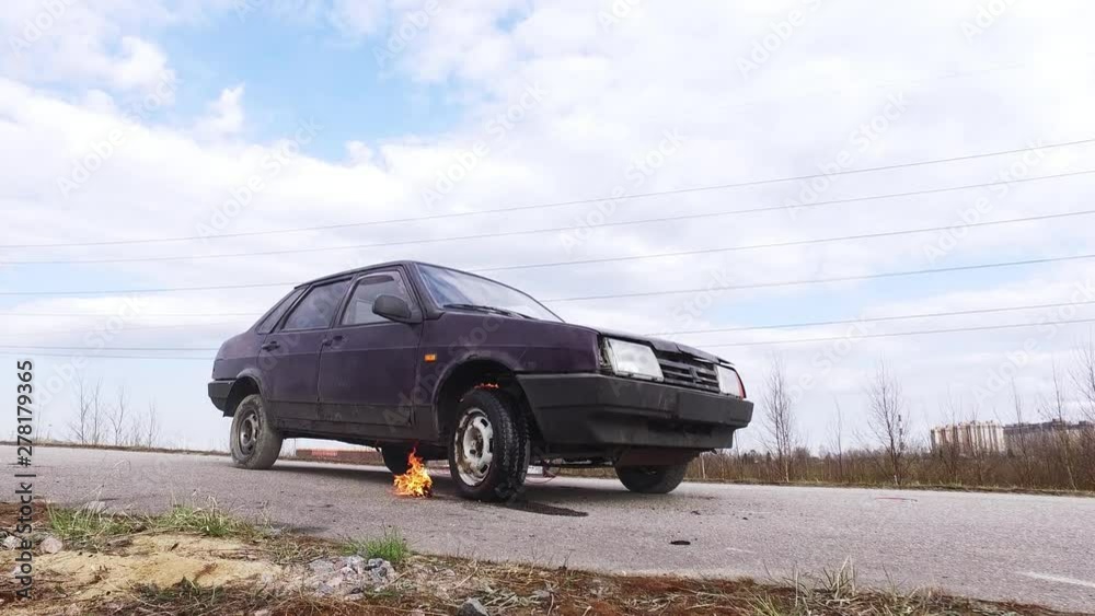 Old dark rusty violet car is left on abandoned road at some suburb area where car's wheel being put on fire on cold winter day. Old dark rusty violet car is left on abandoned road at some suburb area where car's wheel being put on fire on cold winter day.