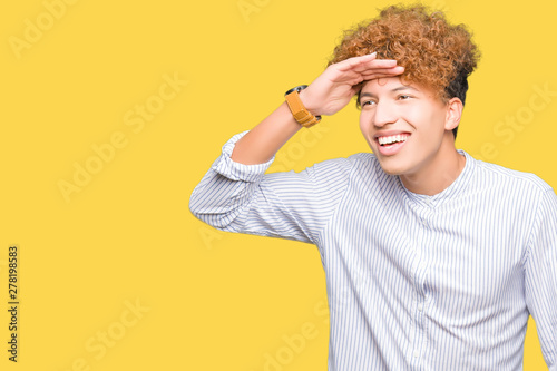 Young handsome business man with afro hair wearing elegant shirt very happy and smiling looking far away with hand over head. Searching concept.