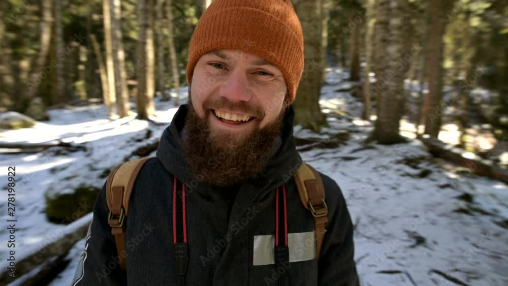Portrait of a cheerful bearded laughing young man with a backpack standing in a coniferous winter forest