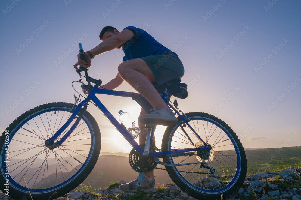 Strong fit male mountain biker performing stunts on rocky terrain on a sunset while wearing a blue shirt and riding a blue bike Strong fit male mountain biker performing stunts on rocky terrain on a sunset while wearing a blue shirt and riding a blue bike