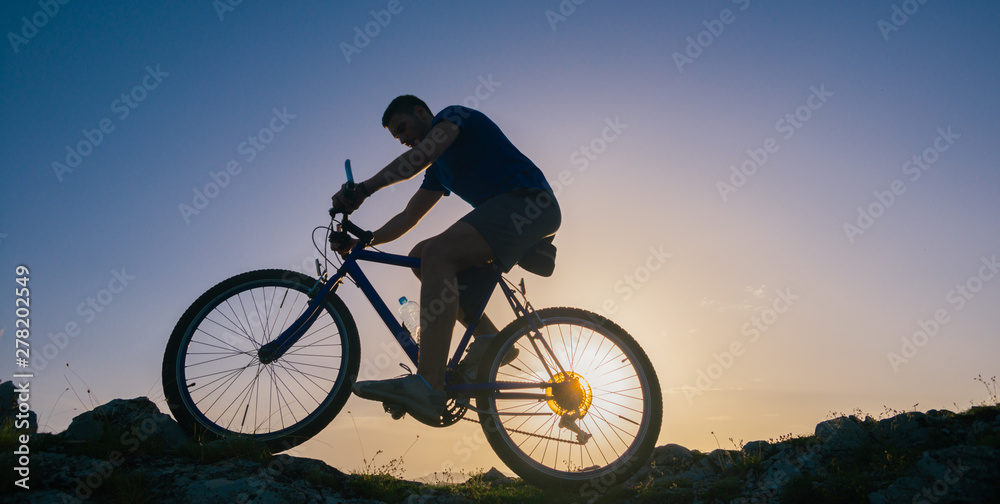 Obraz premium Silhouette of a fit male mountain biker riding his bike uphill on rocky harsh terrain on a sunset.