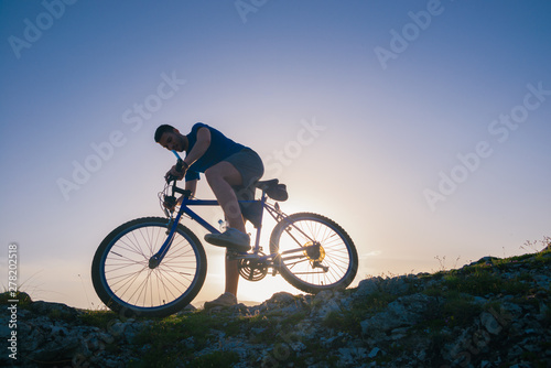 Fototapeta Naklejka Na Ścianę i Meble -  Strong fit male mountain biker performing stunts on rocky terrain on a sunset while wearing a blue shirt and riding a blue bike