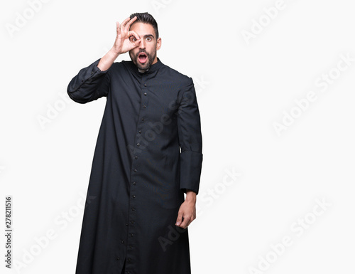 Young Christian priest over isolated background doing ok gesture shocked with surprised face, eye looking through fingers. Unbelieving expression.
