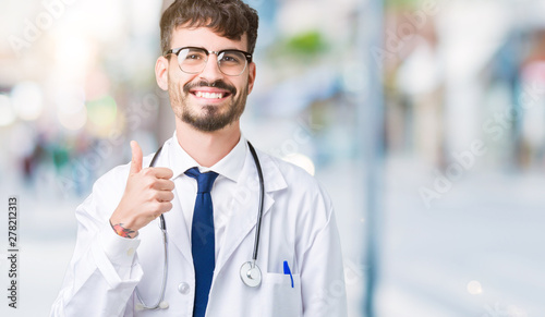 Young doctor man wearing hospital coat over isolated background doing happy thumbs up gesture with hand. Approving expression looking at the camera showing success.