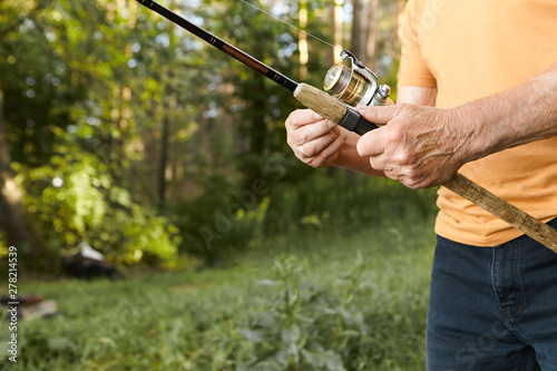 Close up image of elderly man’s hands with wrinkles holding fishing rod. Cropped picture of unrecognizable senior mature male fishing on river bank, standing against green trees background
