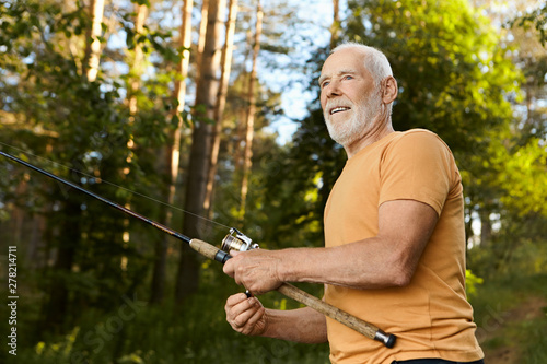 Low angle view of handsome elderly 60 year old male with thick gray beard having joyful facial expression, pulling fish out of water while fishing at lake, spending summer morning outdoors