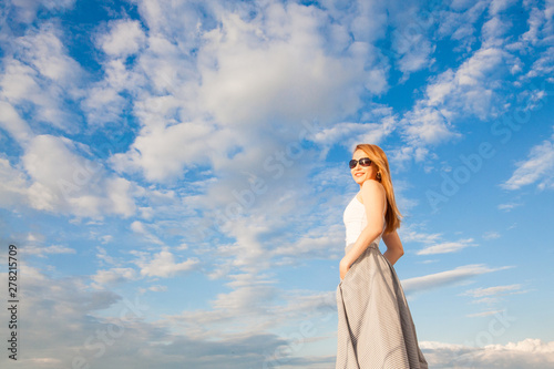 A model in a white T-shirt and long skirt, with long hair, wearing glasses, stands on a white yacht, next to a sail, against a blue sky with clouds, with space for an inscription. Concept vacation.