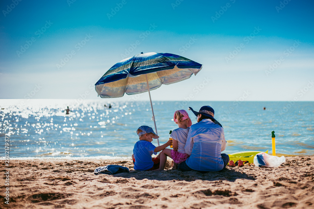 mother with children sit under an umbrella on the seashore on a hot day - obrazy, fototapety, plakaty 