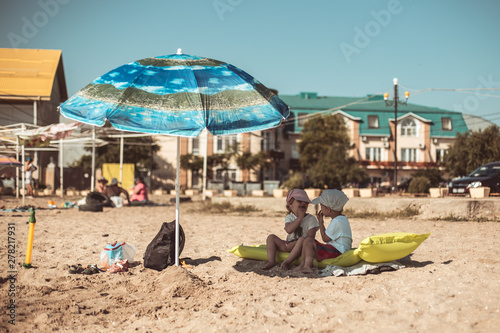 brother and sister sit under an umbrella on the beach on a hot day © Dmitrii