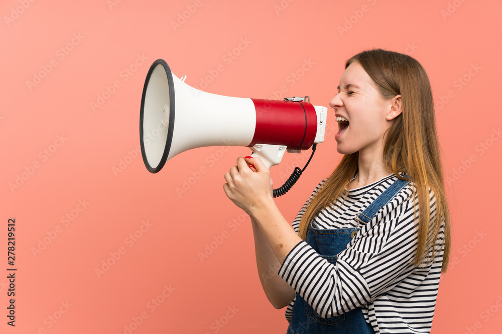 Young woman with overalls over pink wall shouting through a megaphone Young woman with overalls over pink wall shouting through a megaphone