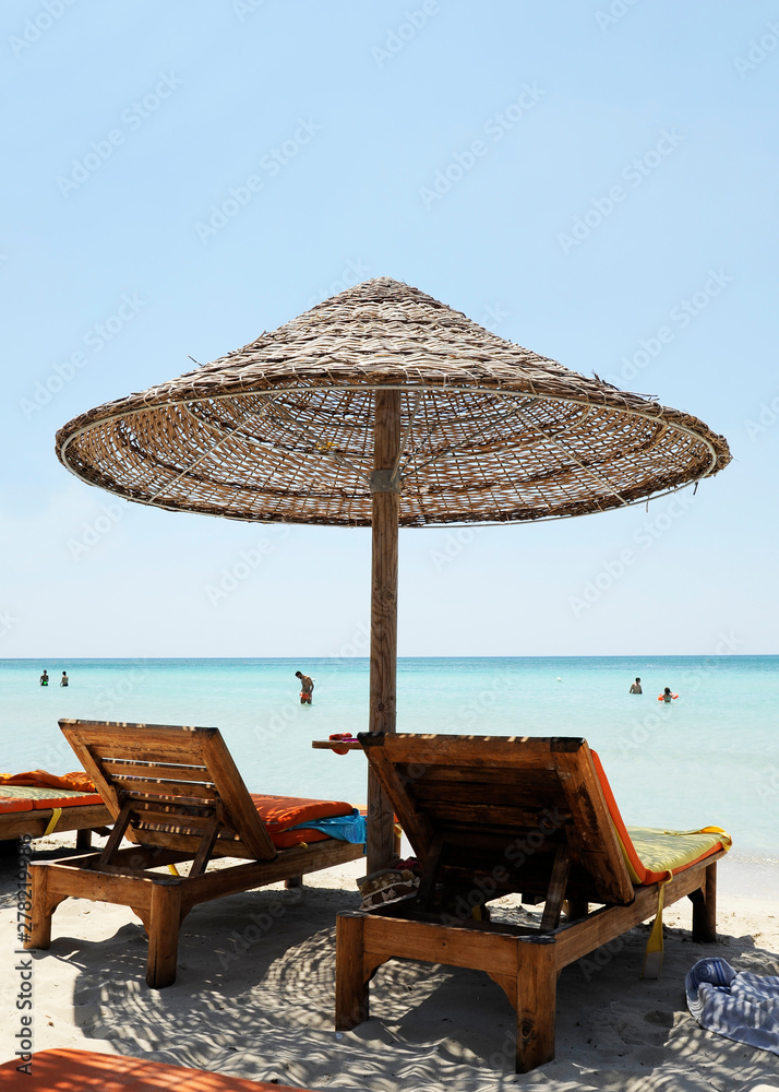 Beautiful beach resort with straw umbrellas on a blue sky and white clouds. On the background some people having fun on the beach. Clear sea. Beach life and lifestyle concept. 