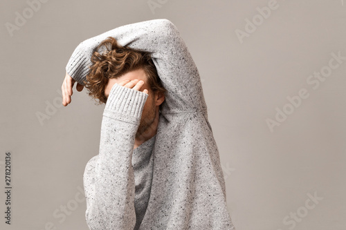 Horizontal studio shot of unrecognizable young male with trendy voluminous hairdo standing in relaxed pose covering his face, dressed in gray knitted sweater. Men’s wear, style and fashion concept