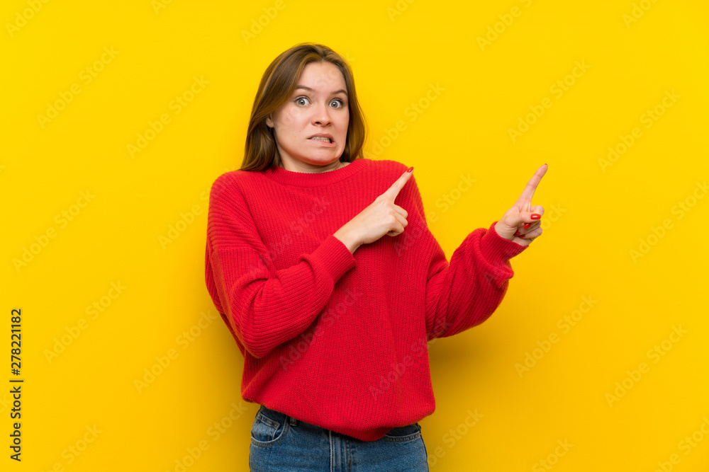 Young woman over yellow wall frightened and pointing to the side Young woman over yellow wall frightened and pointing to the side