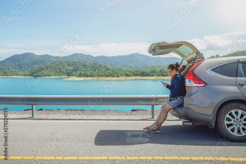woman alone traveler on the road with sitting on hatchback car and playing smartphone with mountain and lake background , concept travel on holiday.
