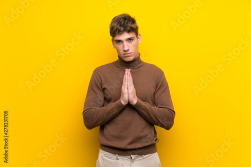 Handsome young man over isolated yellow background keeps palm together. Person asks for something