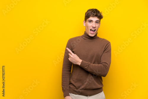 Handsome young man over isolated yellow background surprised and pointing side