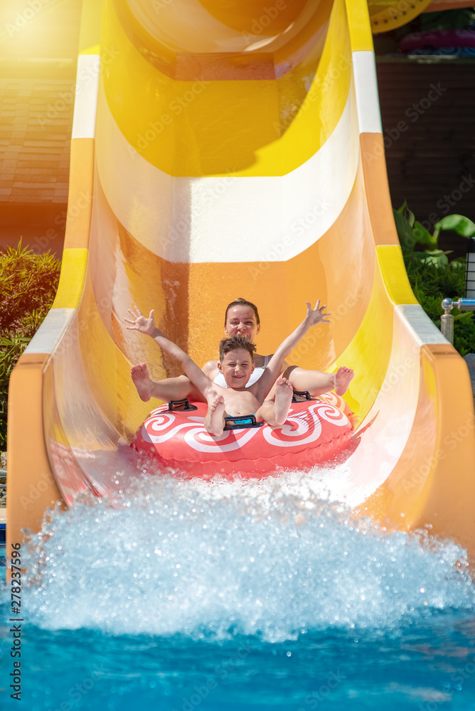 European boy and mom gliding down slide in waterpark. They enjoy the fun and holding hands wide open.
