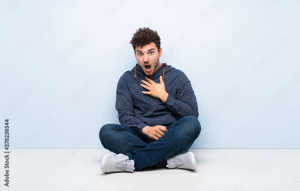 Young man sitting on the floor surprised and shocked while looking right Young man sitting on the floor surprised and shocked while looking right