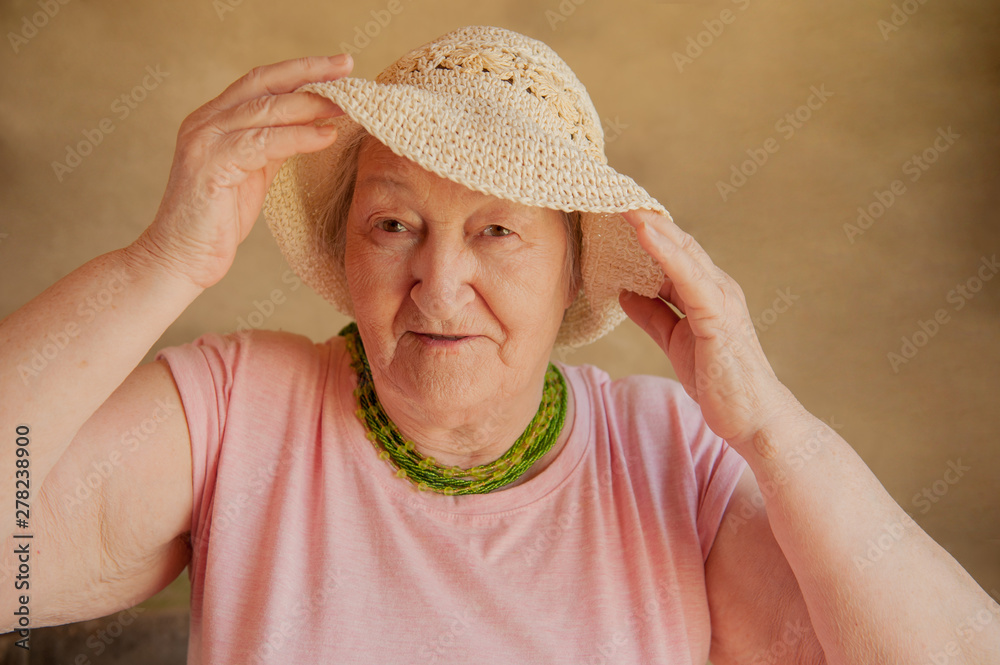 Portrait of elderly woman in pink T-shirt with beads, straw hat and ornaments and glasses. Pensioner supports health and looks great with cosmetic bag