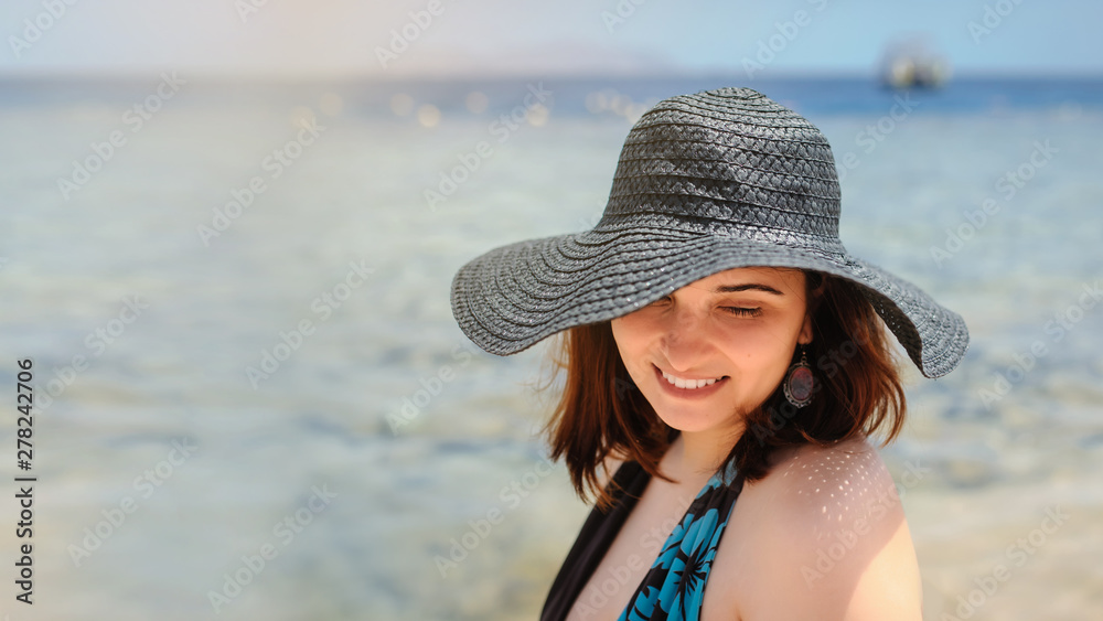 close up photo of a young woman in swimsuit and a blue big hat smiling and sunbathing on the beach