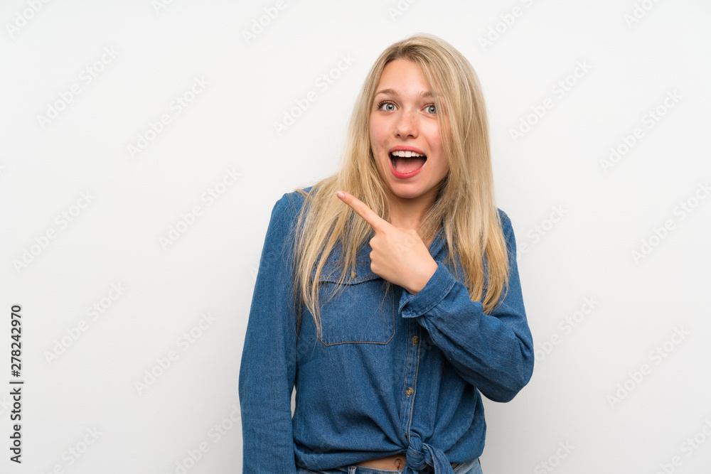 Young blonde woman over isolated white wall surprised and pointing side Young blonde woman over isolated white wall surprised and pointing side